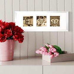 E ♥ M, drei Alphabet Fotos, die die zwei Initiale E für Elli und M für Martin mit einem Fotobuchstaben mit einem Herz verbunden ist. Die Alphabet Fotos sind sepia Farben. Gerahmt sind sie in einem weißen Holzrahmen mit weißem Passepartout. Das individuelle Liebesgeschenk hängt an der Wand über einer weißen Kommode, auf der sich links im Bild rote Blumen in einer rosa Vase und daneben rosa Tulpen befinden.